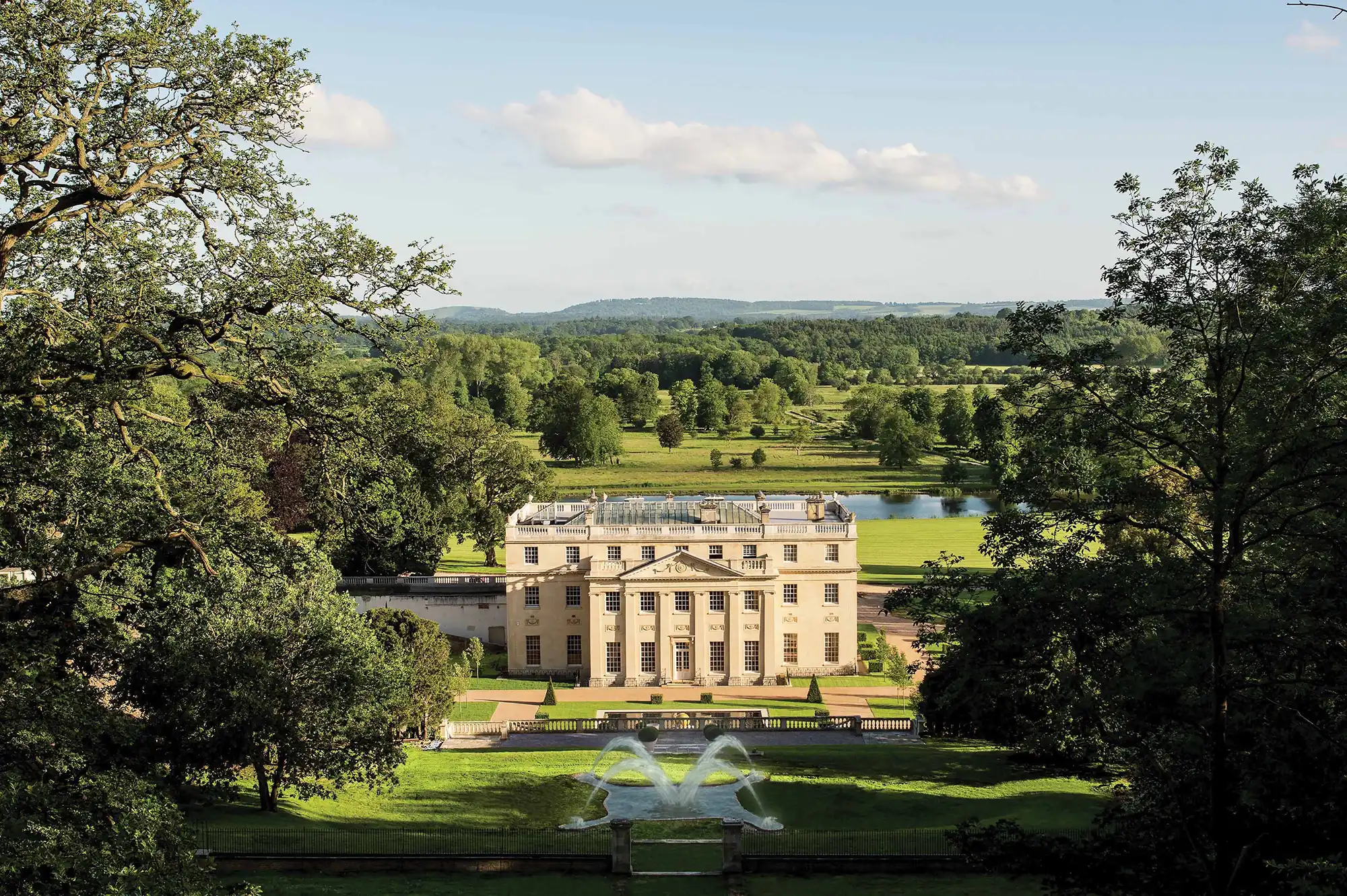 Benham Park, a Georgian Palladian country house set within landscaped parkland, viewed from an elevated position with formal gardens, mature trees, and rolling countryside beyond.
